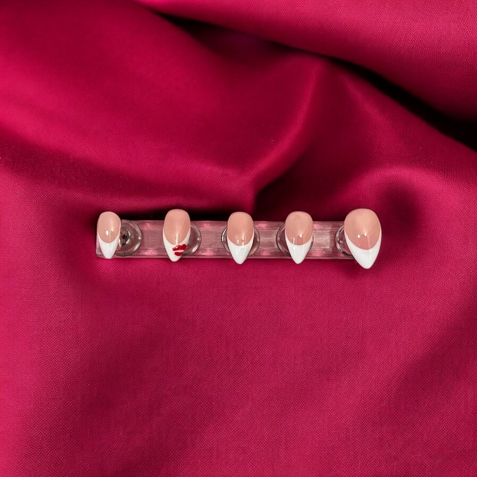 Set of artificial nails with white tips and kiss design on deep pink fabric background.
